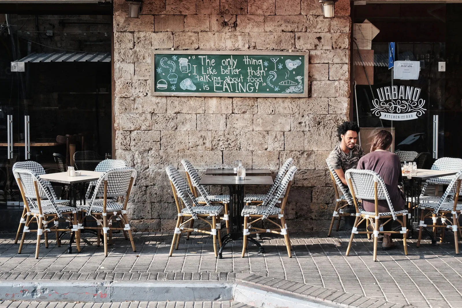 שולחנות וכיסאות של בית קפה על המדרכה לצד קיר אבן, ישיבה ארוכה באמצע היום