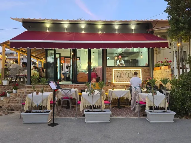 חזית מסעדה בשעת בין ערביים עם סוכך אדום, שולחנות במדרכה ועציצים
