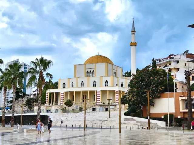 מסגד לבן עם כיפה מוזהבת וצריח גבוה, דקלים וכיכר רטובה לפניו