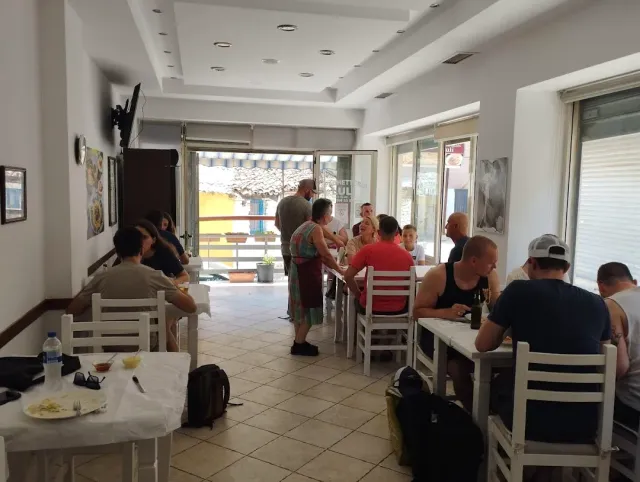 חדר בהיר עם כיסאות פלסטיק לבנים וסועדים סביב שולחנות, ודלת פתוחה לרחוב