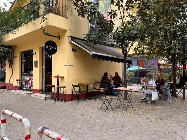 מבנה פינתי צהוב עם שלט קטן, שולחנות בחוץ ויושבים תחת העצים