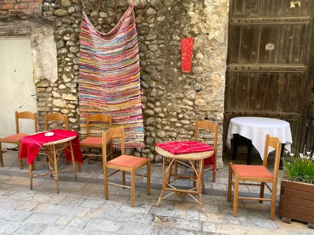שטיח סמרטוטים צבעוני תלוי על קיר אבן, ולפניו שולחנות קטנים וכיסאות עץ
