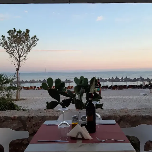 שולחן במפה ורודה עם בקבוק יין, ומולו חוף עם שמשיות קש וים בדמדומים