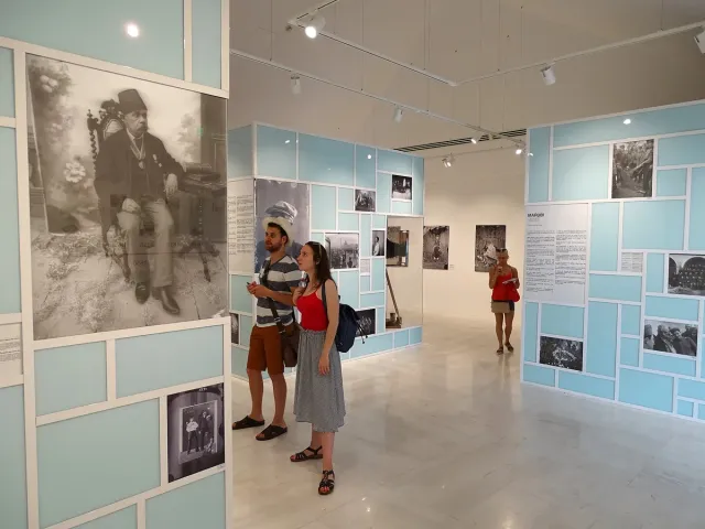 אולם תצוגה בהיר עם צילומים היסטוריים על קירות תכלת, ומבקרים מתבוננים בדיוקן מוגדל
