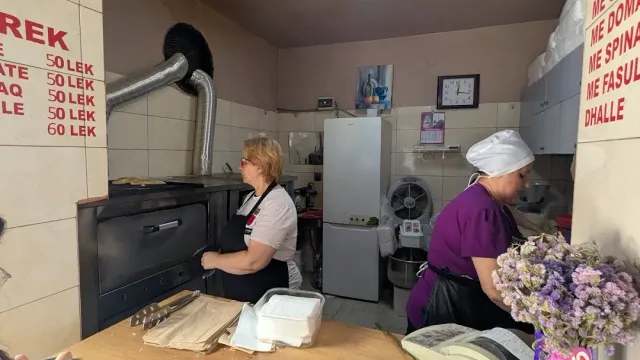 שתי עובדות ליד תנור מתכת במאפייה, מחירון על הקיר ומגשים על הדלפק