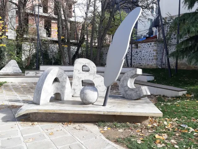 פסל אבן בצורת אותיות ונוצת כתיבה בחצר מרוצפת עם עצים חשופים