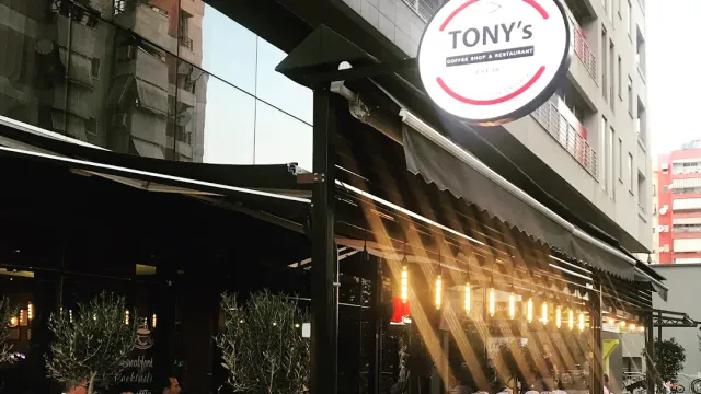 שלט עגול מעל סוכך מרפסת, שרשרת נורות ועצי זית בעציצים בערב