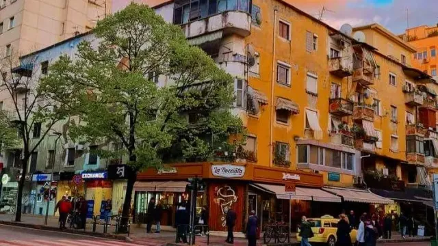 רחוב עם בנייני מגורים צבעוניים, עצים ירוקים וחנות פינתית עם סוכך