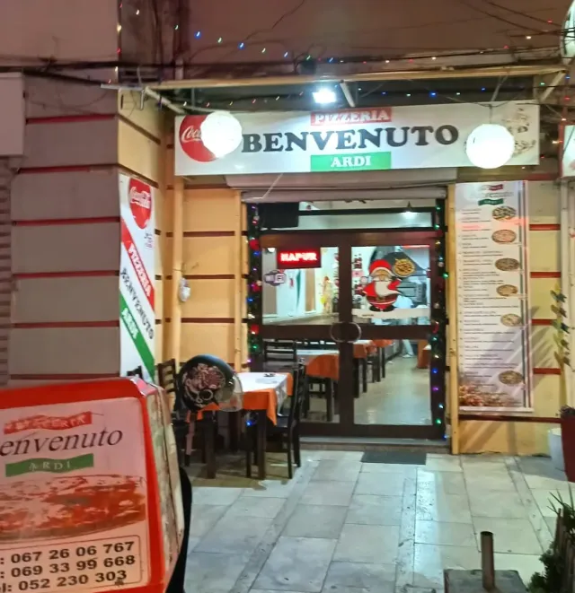 כניסה מוארת בלילה עם שלט לבן, נורות צבעוניות ושולחנות במפות כתומות בפנים