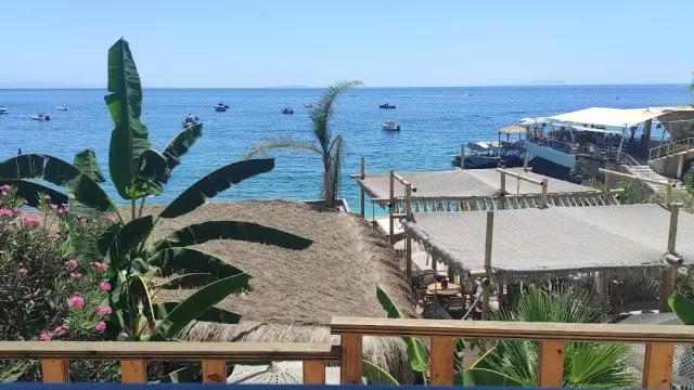 מבט ממרפסת עץ על גגות קש של חוף, עלי בננה וים כחול עם סירות