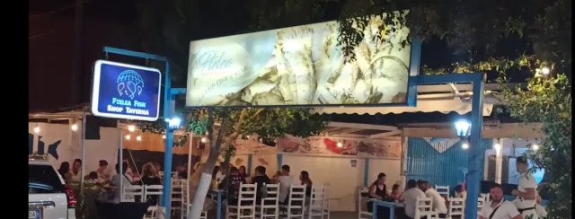 טברנה בלילה עם שלטים מוארים, כיסאות לבנים וסועדים תחת נורות תלויות