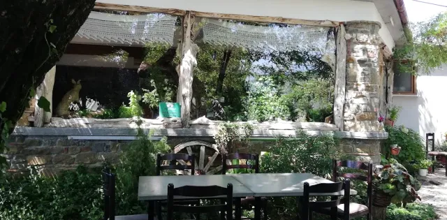 גינה מוצלת עם שולחנות וכיסאות, קיר אבן, רשת צל וגלגל עגלה ישן