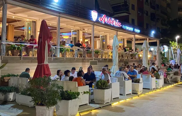 מרפסת הומה סועדים בלילה עם שלט מואר, שמשיות סגורות ואדניות לבנות