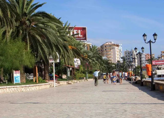 טיילת מרוצפת רחבה בין דקלים גבוהים ופנסי רחוב, עם הולכי רגל ובנייני נופש