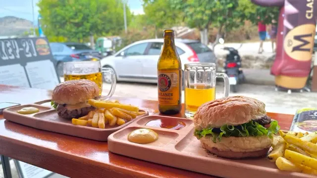 שני המבורגרים על מגשי עץ עם צ׳יפס, בקבוק בירה וכוסות על שולחן חוץ