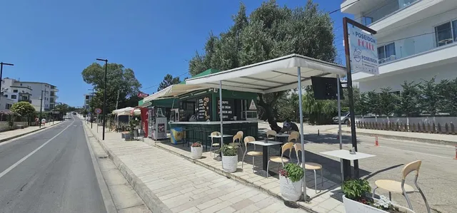 קיוסק ירוק עם סוכך לבן ושולחנות קטנים על מדרכה רחבה ליד הכביש